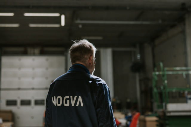 Mann med ryggen til iført mørk arbeidsjakke med NOGVA-logo i et dempet industrirom med verkstedsutstyr i bakgrunnen.