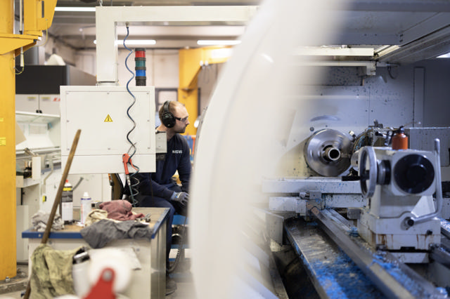 Ansatt med hørselsvern og vernebriller overvåker en dreiebenk i drift på mekanisk verksted, omgitt av verktøy og maskindeler.